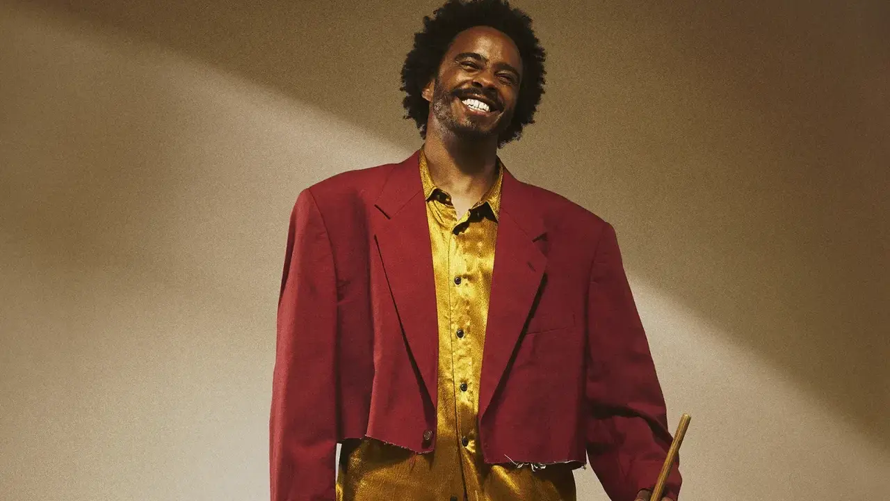 Un percussionniste célèbre, souriant, porte une veste courte bordeaux et une chemise dorée, tenant une baguette.