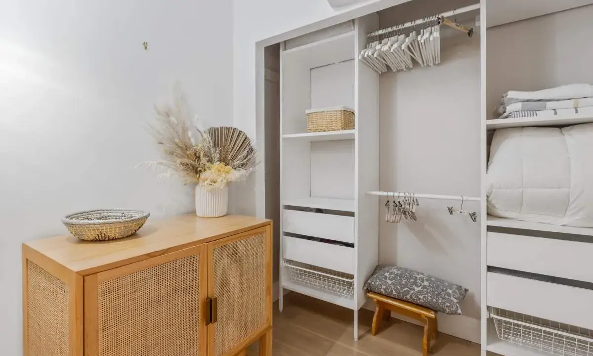 Un armario blanco organizado, listo para limpiar armarios por dentro. Cestas, ropa de cama y un jarrón con flores secas decoran el espacio.