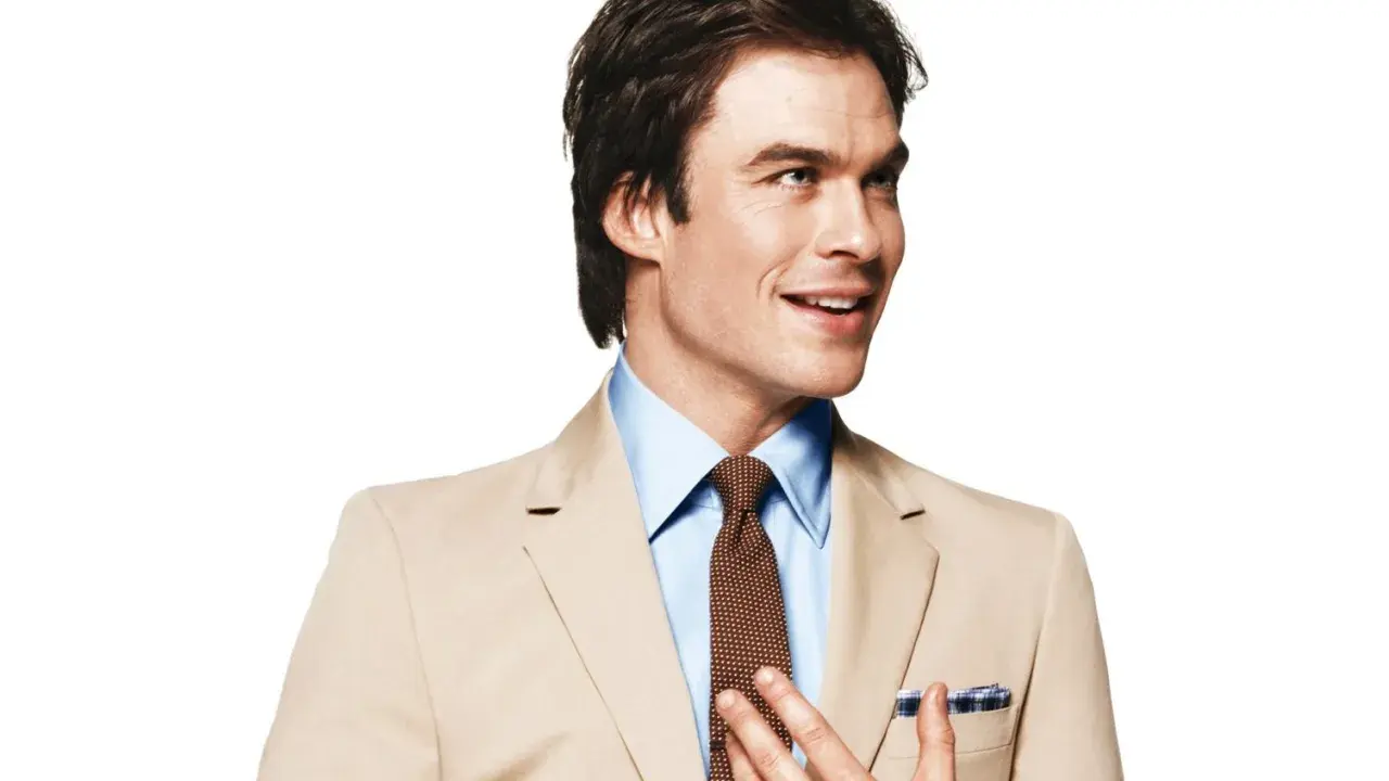A man in a tan suit and blue shirt smiles, adjusting his tie.