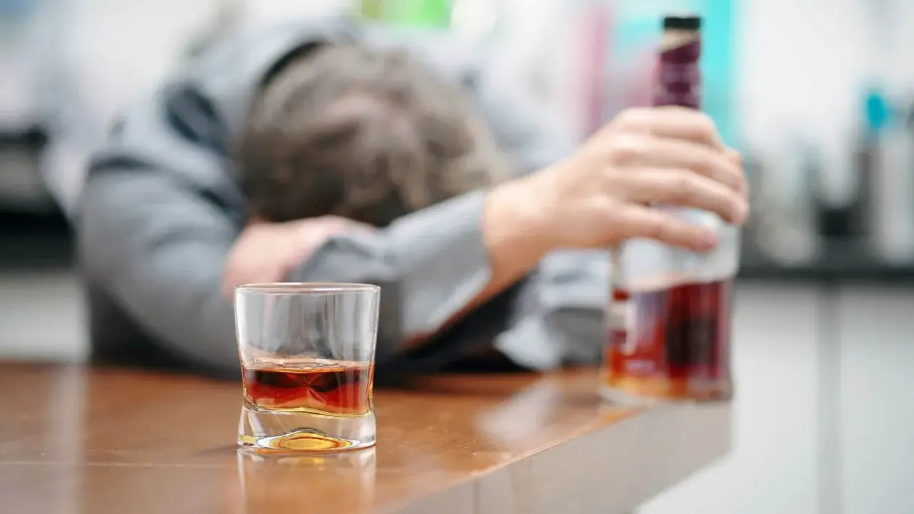 Zaniedbany mężczyzna, prawdopodobnie po eksmisji alkoholika z domu, leży na stole obok szklanki z whisky i butelki.