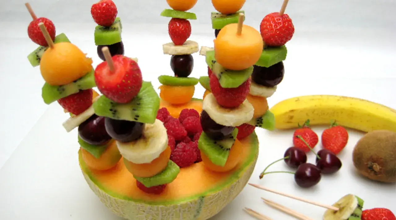 Une **brochette de fruits** colorée avec fraises, kiwis, bananes, cerises et melon, présentée dans une moitié de melon.