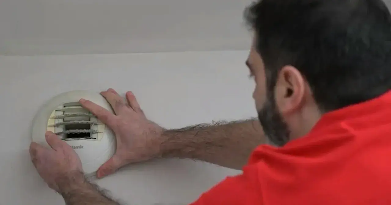 Un homme en chemise rouge effectue l'entretien d'une VMC murale, vérifiant le filtre encrassé.