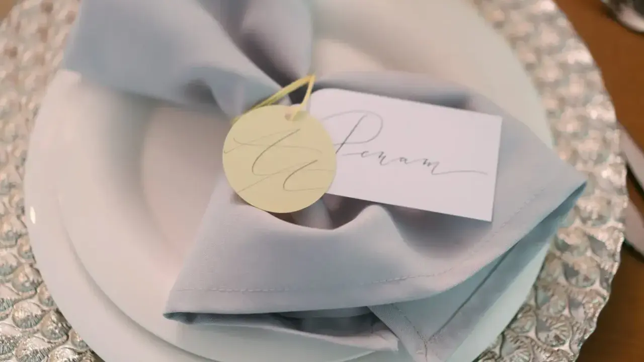 Elegancka winietka z imieniem "Peram" na jasnoszarym obrusie, ozdobiona okrągłym, żółtym tagiem.