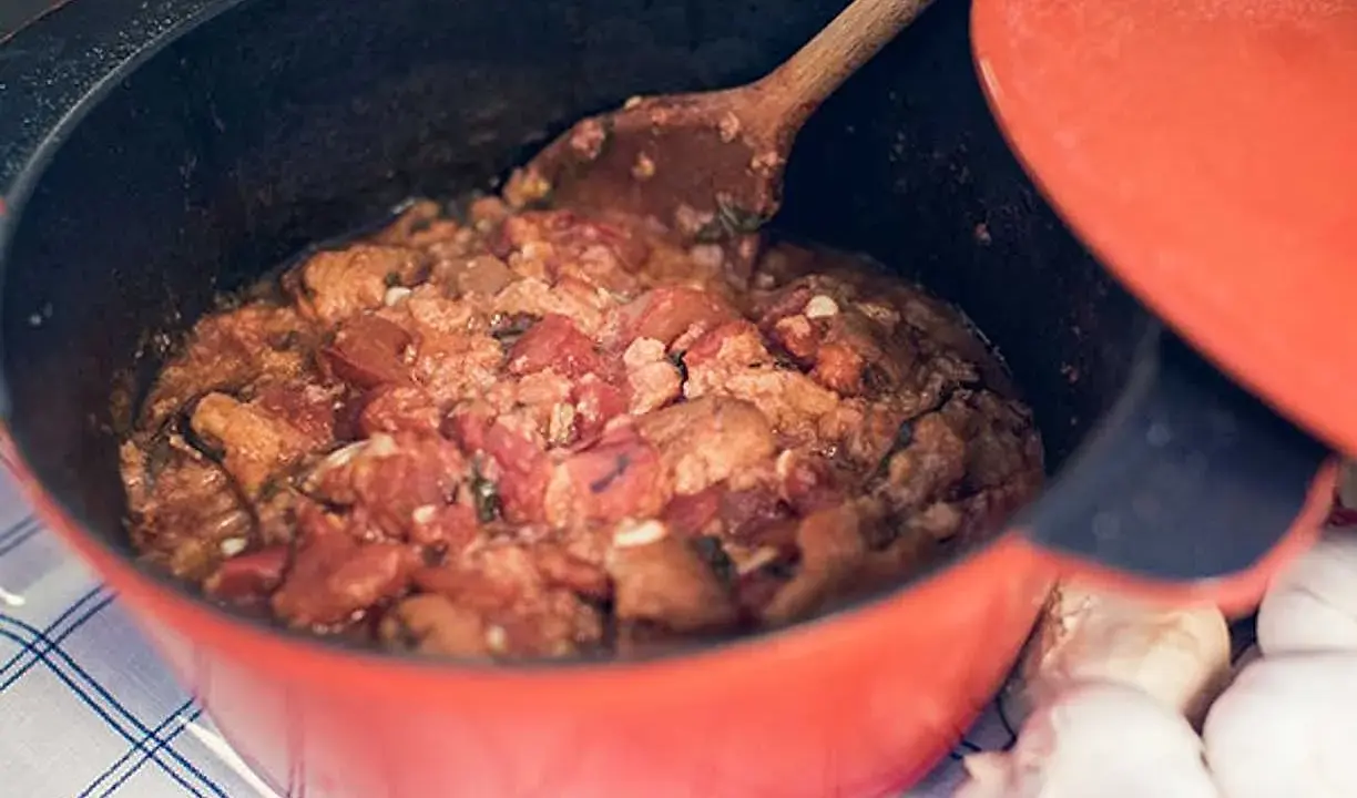 En röd gryta med en mustig toscansk tomatsoppa, en träsked rör om i den väldoftande grytan. Bredvid ligger vitlök.