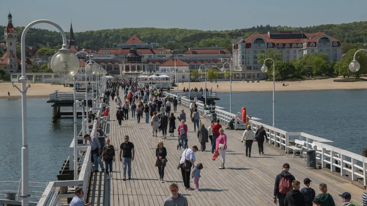 Tłum ludzi spaceruje po molo w Sopocie, jednym z nietypowych atrakcji Gdańska. W tle widać plażę i zabytkowe budynki.