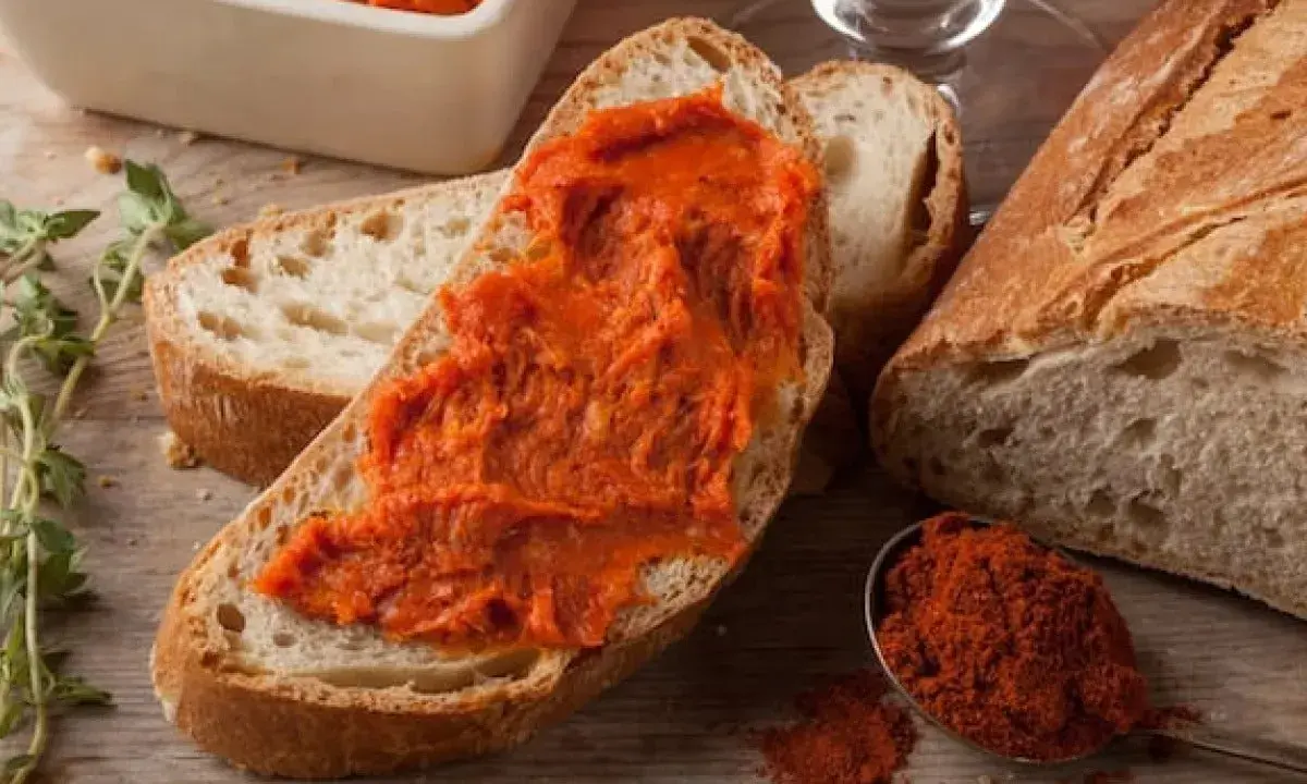 Fetta di pane con 'nduja, un assaggio di cosa mangiare in Calabria. Spezie rosse e pane fresco completano la scena.
