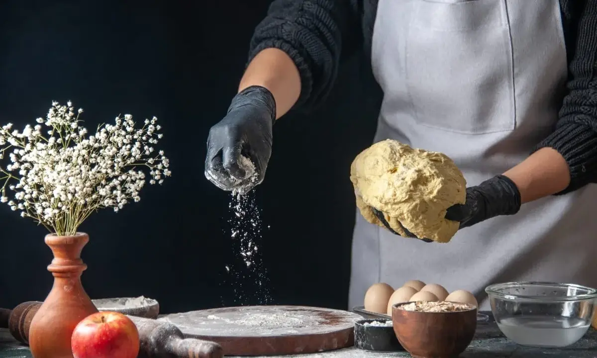 Hände in schwarzen Handschuhen streuen selbsttreibendes Mehl über Teig. Ein Apfel und Blumen sind auf dem Tisch.
