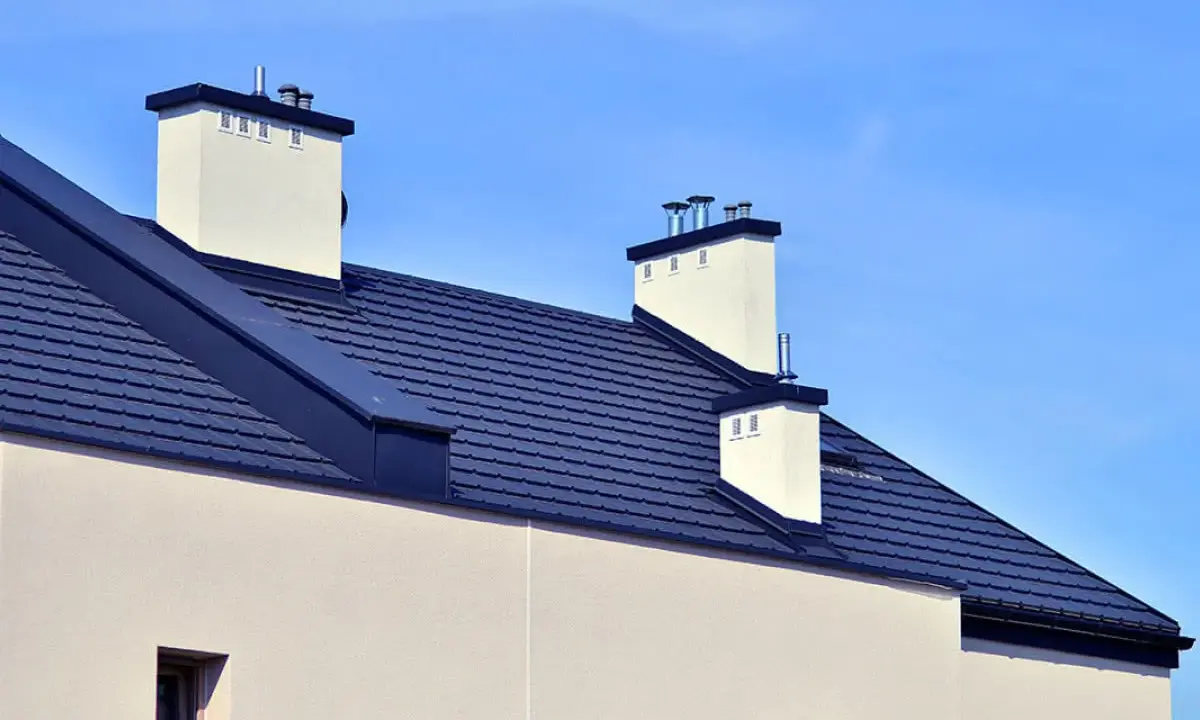 Nowoczesny dach bezokapowy z ciemnogranatową dachówką i białymi kominami, pod błękitnym niebem.