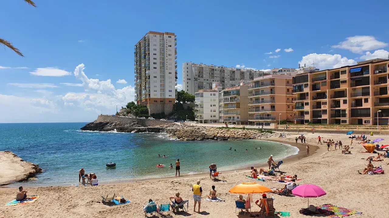 Sonniger Cullera Strand mit vielen Menschen, die sich im Meer und am Sand entspannen, im Hintergrund hohe Gebäude.