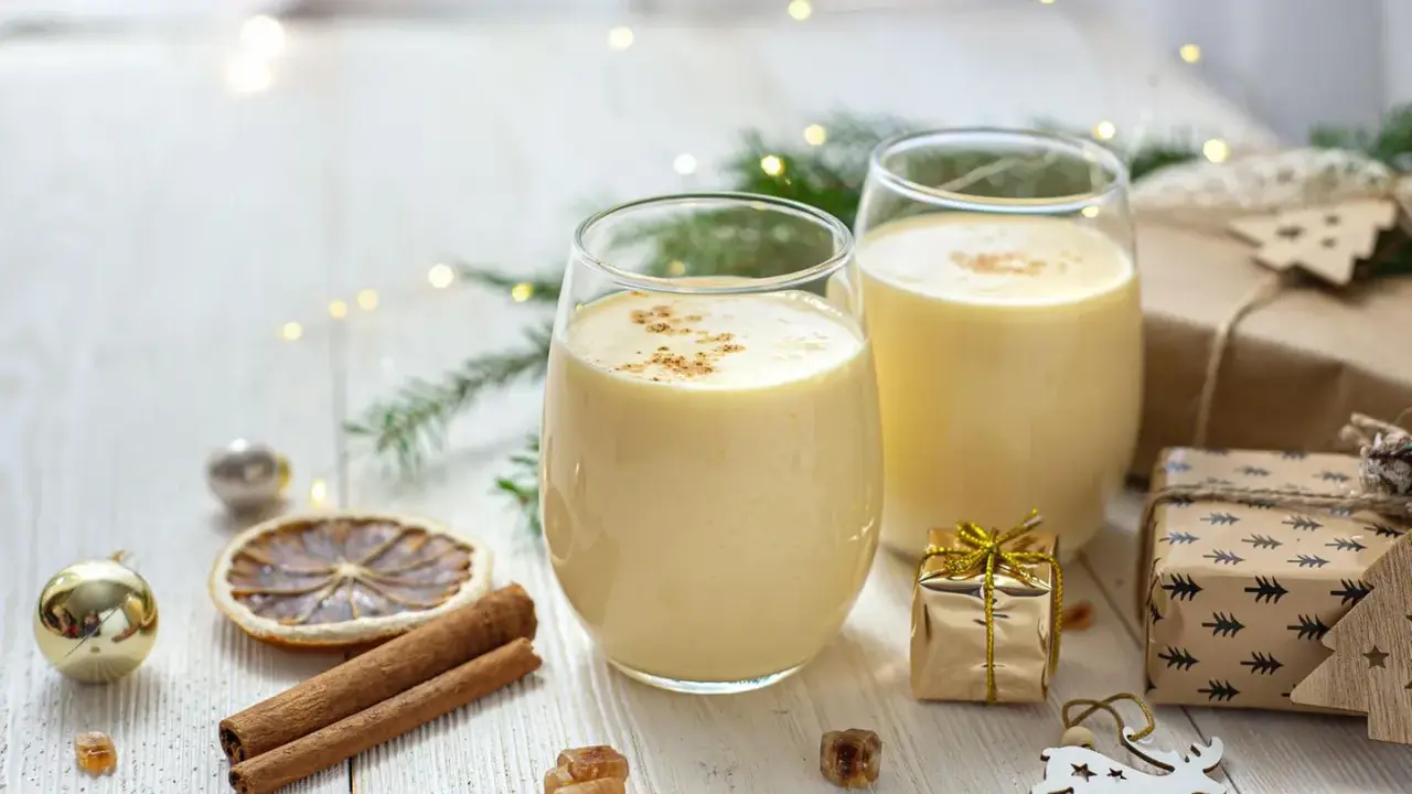 Zwei Gläser cremiger Cocktail mit Eigelb, garniert mit Zimt, umgeben von Weihnachtsdekorationen und Geschenken.