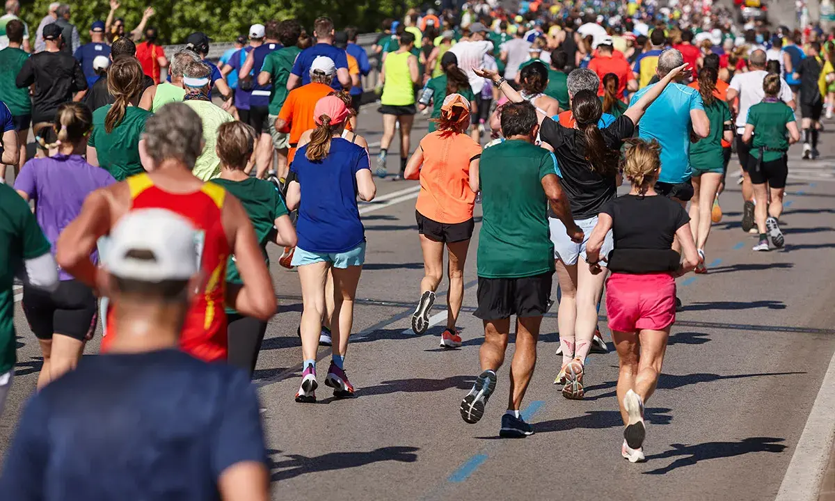 Tłum biegaczy pokonuje kolejne kilometry maratonu. Widać ich nogi w ruchu, kolorowe stroje i cienie na asfalcie.