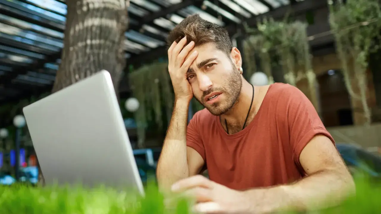 Mężczyzna z ręką na czole, sfrustrowany pracą przy laptopie. Czyżby padł ofiarą pomówienia w internecie?
