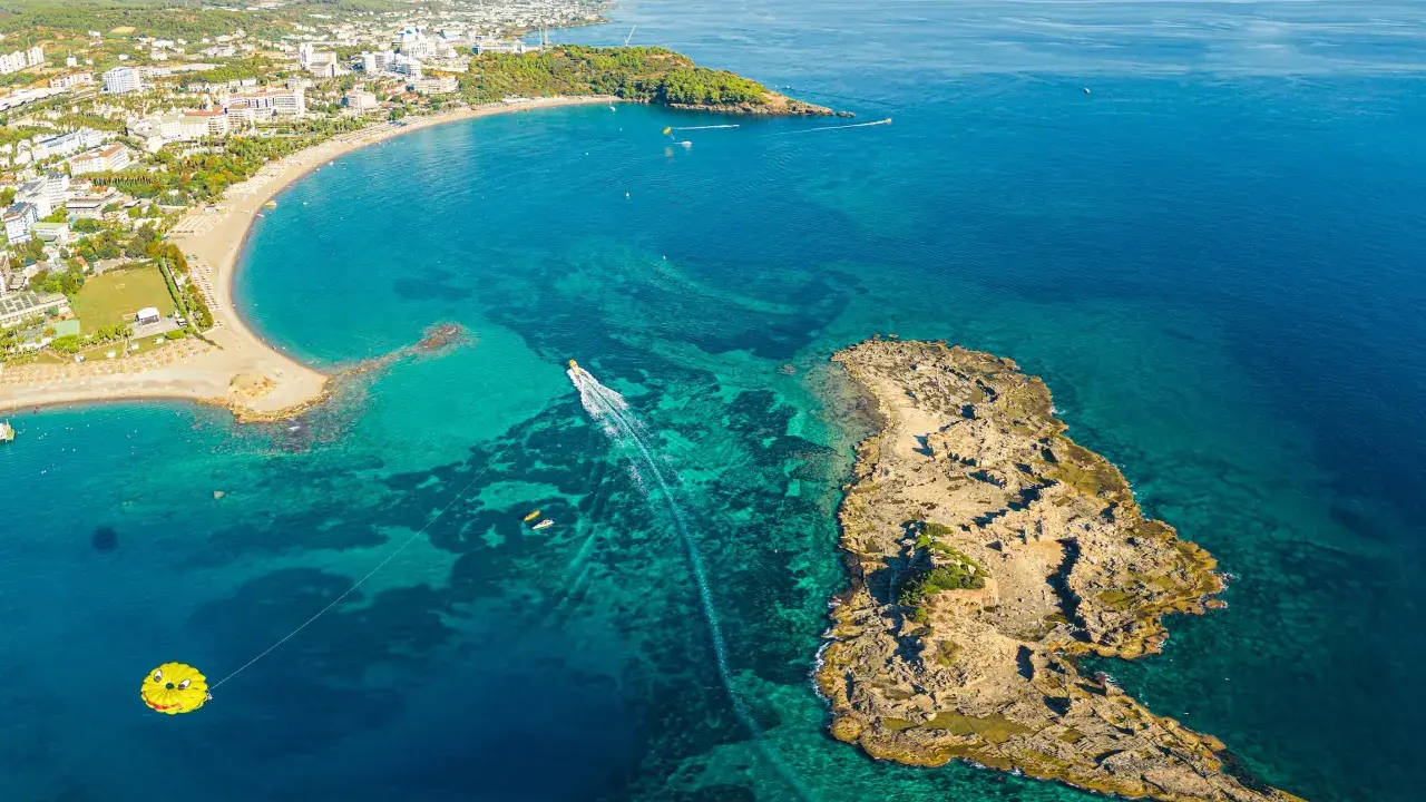 Widok z lotu ptaka na wybrzeże z hotelami, plażą i turkusową wodą. Widać łódź, parasailing i skaliste wysepki, które są świetnymi okurcalar atrakcje.