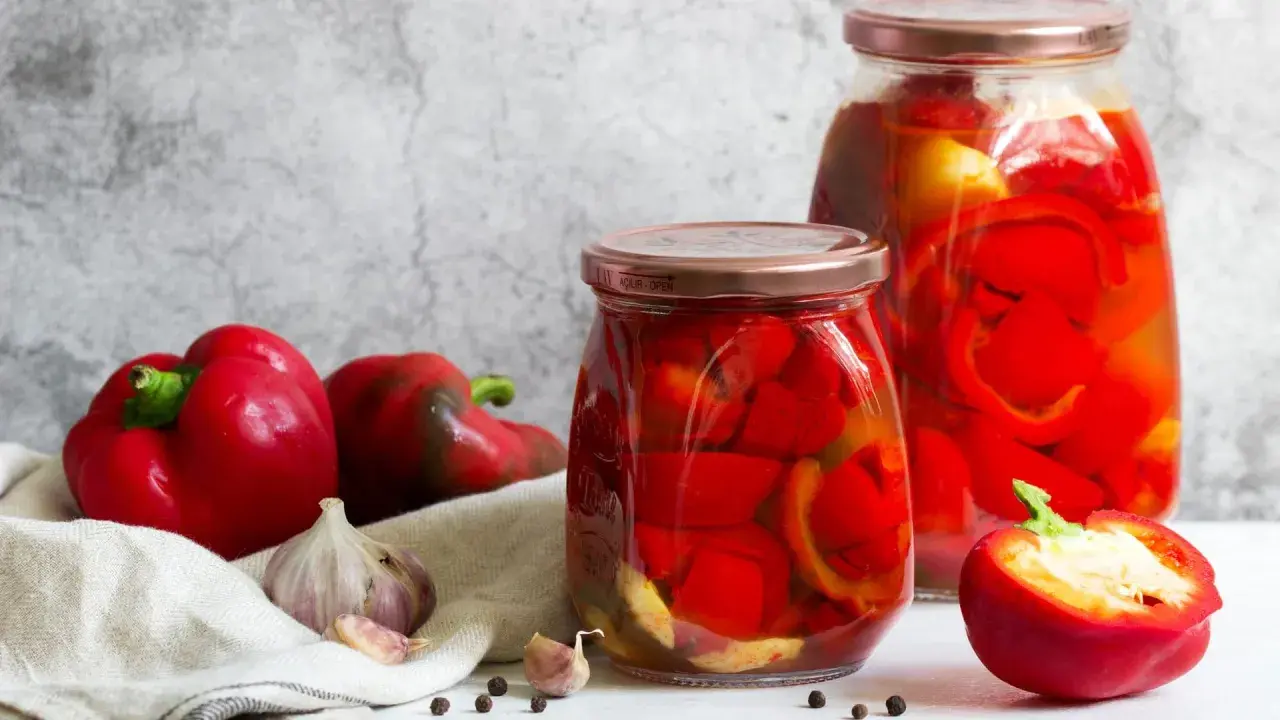 Zwei Gläser mit eingelegter Paprika, daneben frische rote Paprika, Knoblauch und Pfefferkörner. Perfekt zum Paprika einkochen.