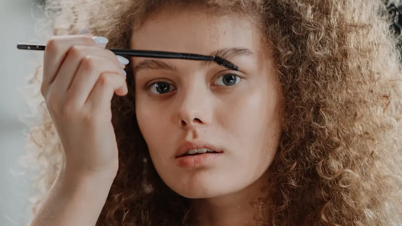 Une jeune femme aux cheveux bouclés applique un produit sur ses sourcils décolorés avec une brosse.