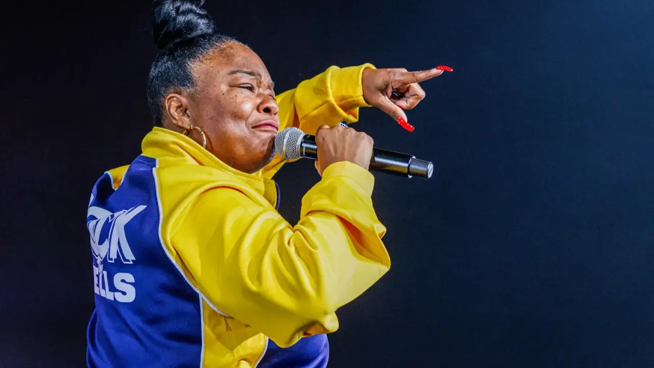 Roxanne Shanté w żółtej kurtce z niebieskimi wstawkami śpiewa do mikrofonu, wskazując palcem.