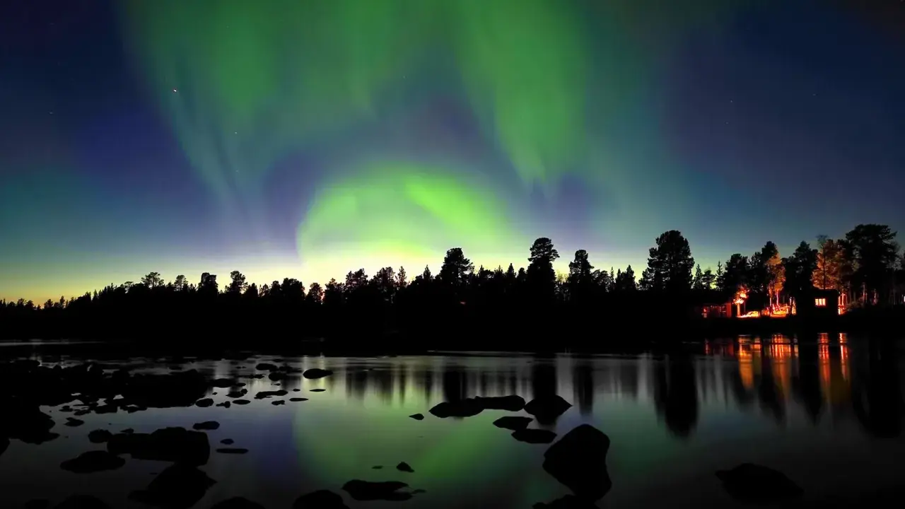 Norrsken dansar över en vinterlandskap vid Kiruna. En skogskant och en sjö med stenar speglar himlens magiska ljus.