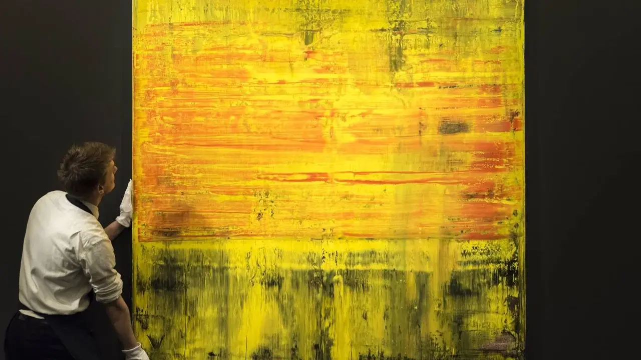 A man in white gloves carefully handles a large abstract painting with vibrant yellow and orange hues, a testament to Gerhard Richter's net worth.