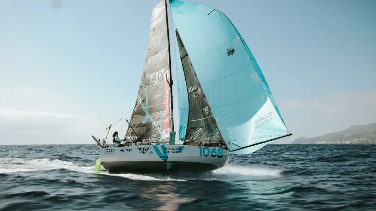 Un voilier de course, le numéro 1068, navigue sur une mer agitée. Les voiles, l'une noire et l'autre bleu clair, portent le nom "la maison perchée". Thais Le Cam est à la barre.