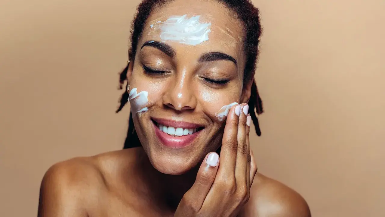 Une femme applique de la crème sur son visage. Elle sourit, les yeux fermés. Elle se demande que veut dire indice 50 pour sa protection solaire.