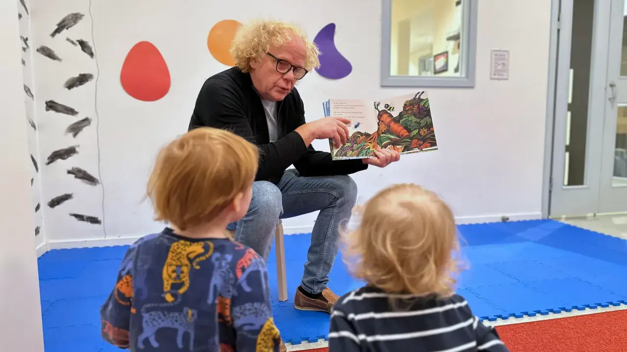 En man med lockigt hår läser en bok för två små barn. Bilden visar en humlan djojj på en morot i boken.