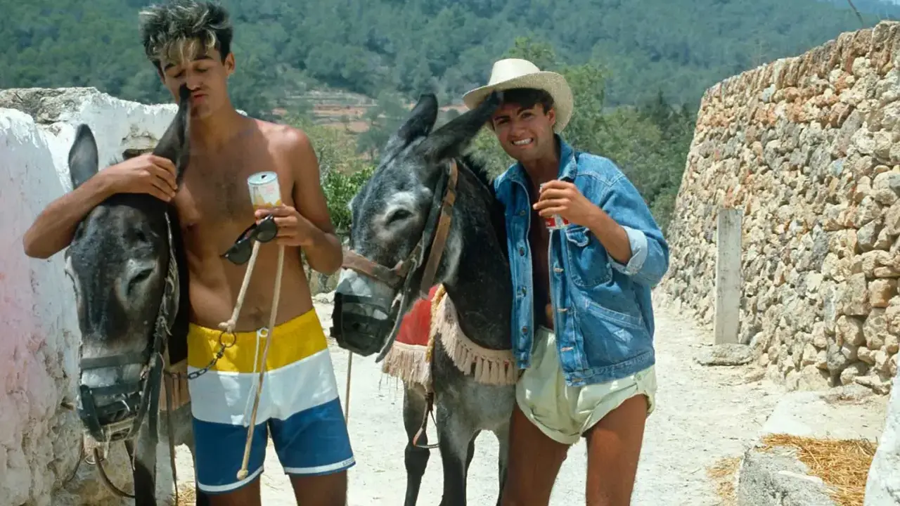 Tony Pike Ibiza: due ragazzi con asini, uno con pantaloncini colorati, l'altro con giacca di jeans e cappello di paglia, sorseggiano bevande.