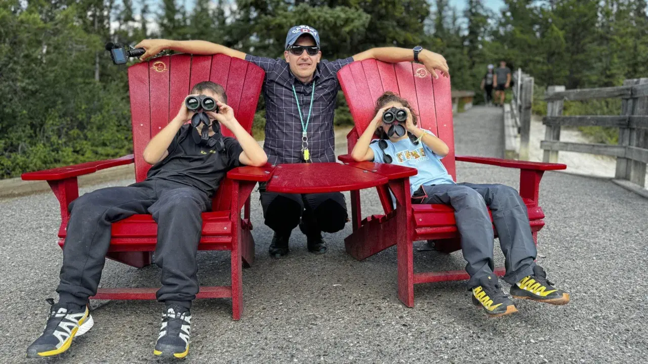 Un padre y sus dos hijos disfrutan de la naturaleza en Canadá con niños, observando el paisaje con binoculares.