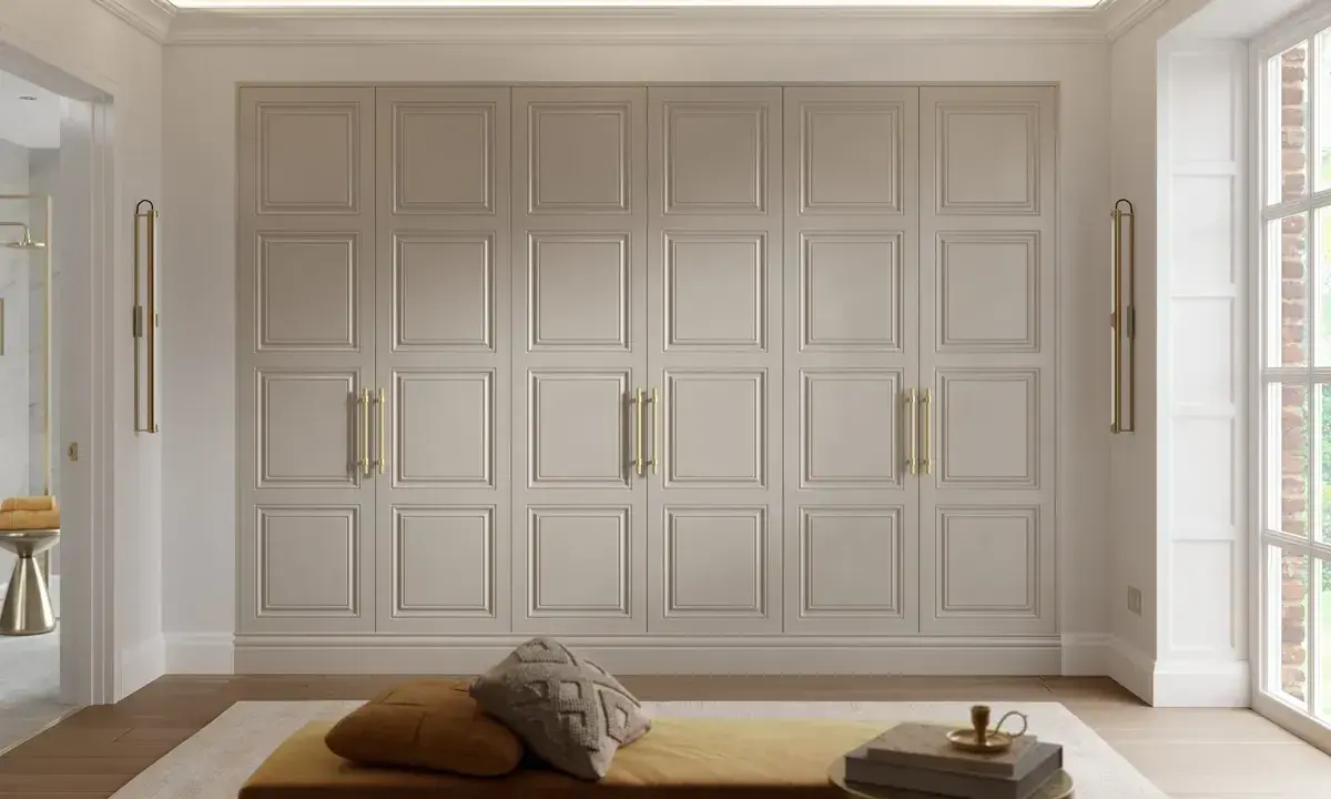 En stor garderob med klassiska paneler och guldfärgade handtag. En soffa med kuddar och en bokhylla framför. En inredning som utstrålar personlig stil.