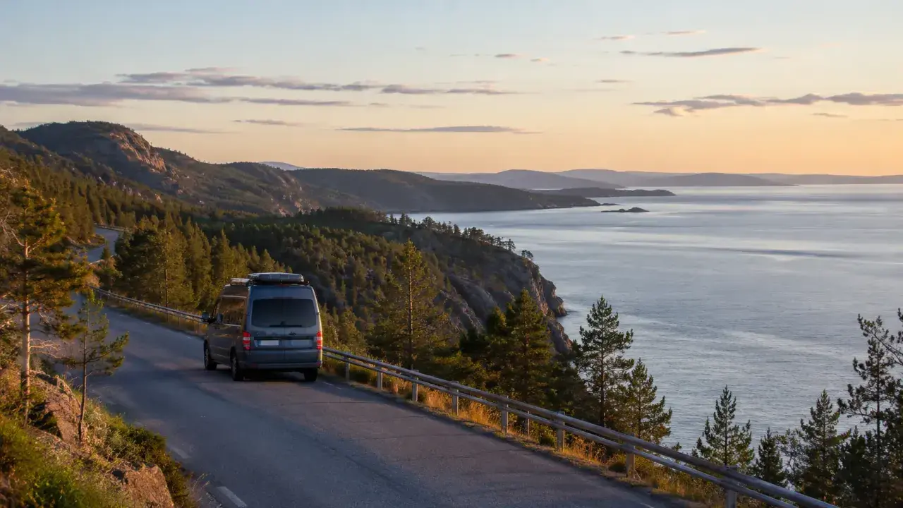En husbil kör längs en slingrande väg vid den vackra Höga kusten. Solnedgången färgar himlen i mjuka toner över det glittrande havet.