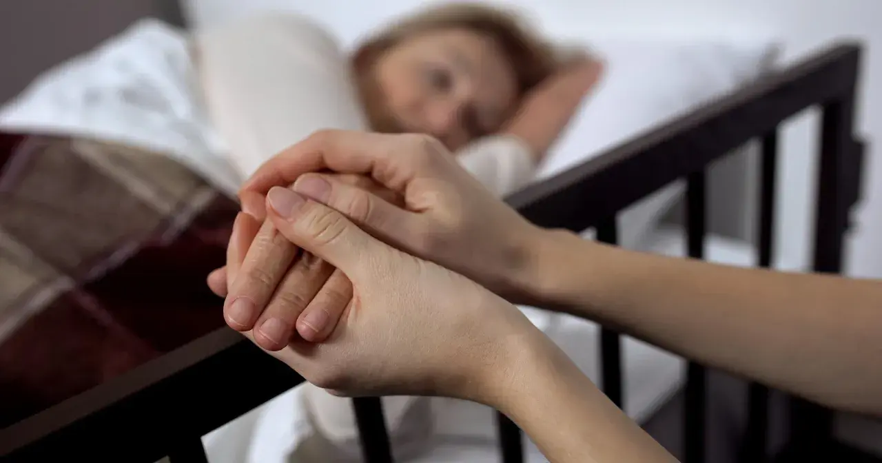 Dwie dłonie splatają się, jedna z nich spoczywa na ramieniu śpiącej osoby. To obraz towarzyszenia w trudnych chwilach, gdy rozważana jest eutanazja co to.