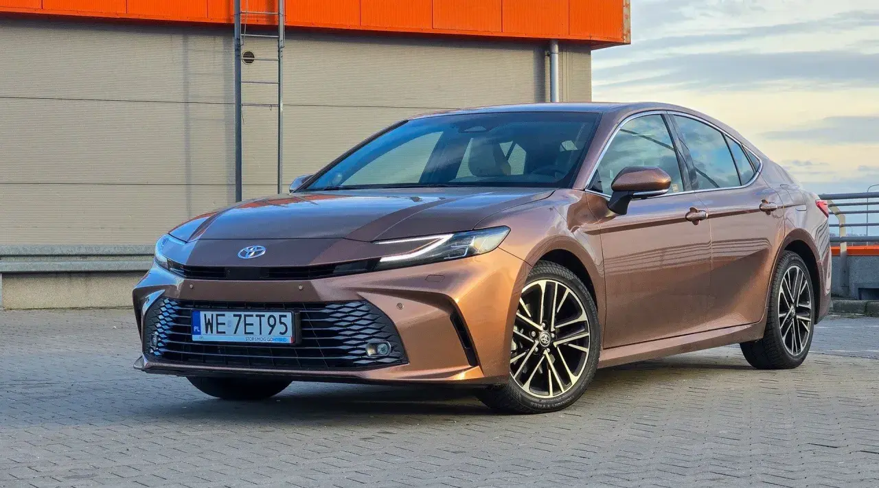 Brązowa Toyota Camry 2.5 Hybrid z eleganckimi felgami stoi na tle nowoczesnego budynku.