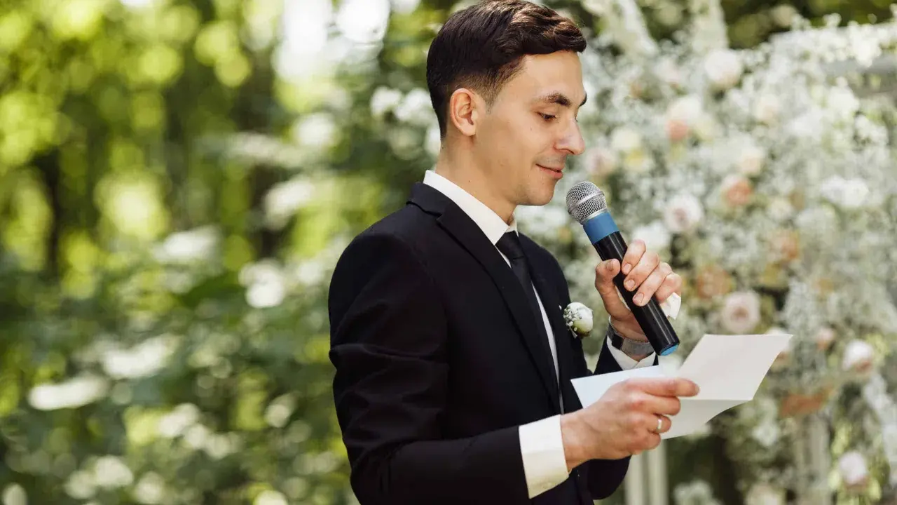 El maestro de ceremonias, con traje negro y micrófono, lee un discurso frente a un arco de flores.