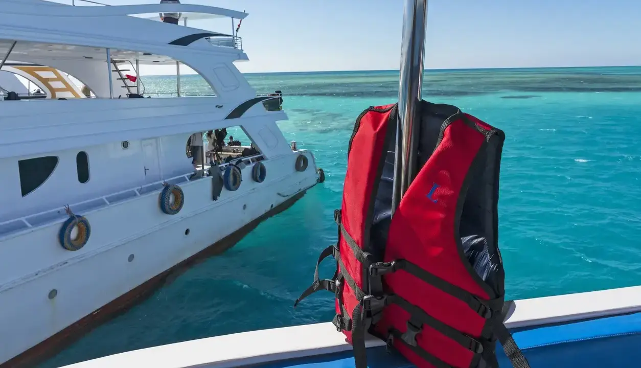 Gilet de sauvetage rouge prêt pour l'aventure en mer, à côté d'un yacht blanc. Les **œuvres mortes du bateau** se reflètent dans l'eau turquoise.