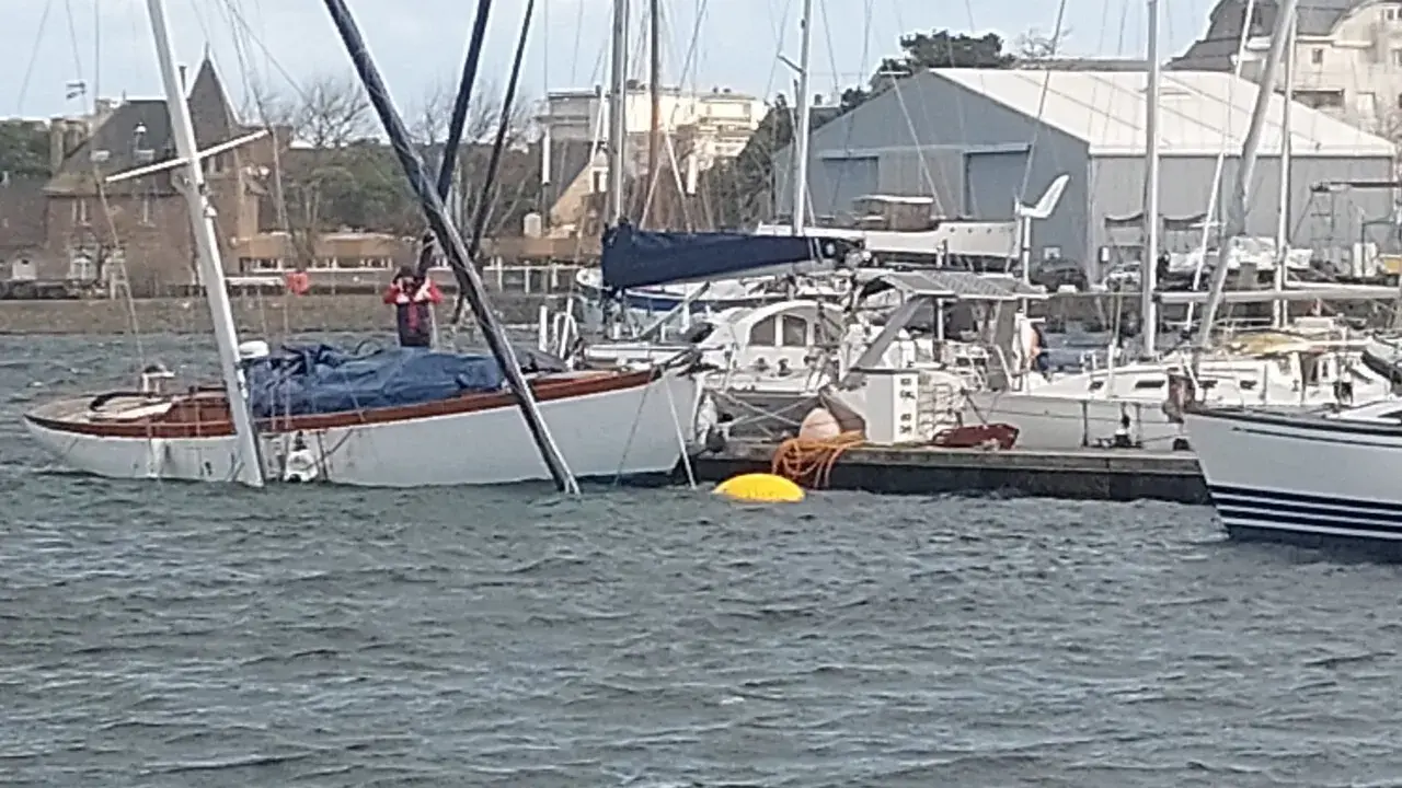 Un voilier blanc et bois, bâché, est partiellement immergé dans un port. Un homme en gilet rouge est à bord.