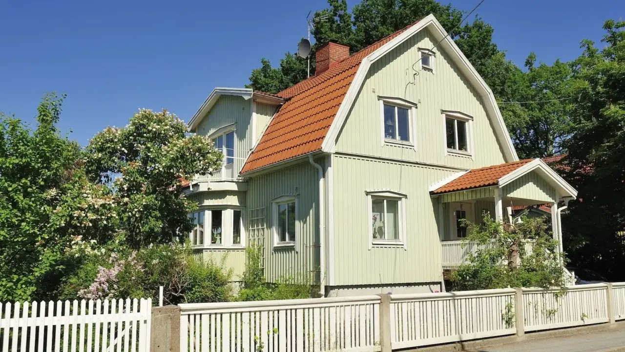 Ljusgrönt trähus med tegeltak och vit staket. En vacker trädgård med blommande buskar. Planerar att byta fasad för att fräscha upp huset.
