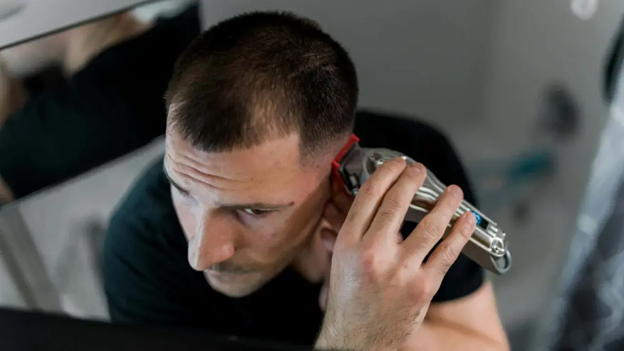 Mann mit Haarschneidemaschine am Hinterkopf. Er schneidet sich die Haare selber, wie eine Frau, die ihre Haare zu Hause schneidet.