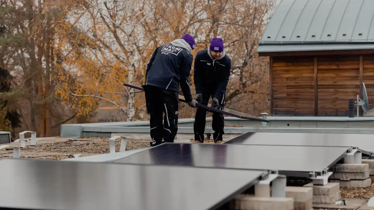Två personer installerar solceller på ett papptak. Höstfärgade träd syns i bakgrunden.