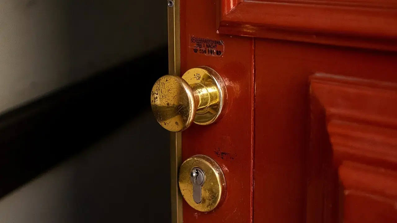 Poignée et serrure dorées sur une porte rouge. Une étiquette indique un service de serrurerie, utile en cas de porte forcée, pour l'assurance propriétaire ou locataire.