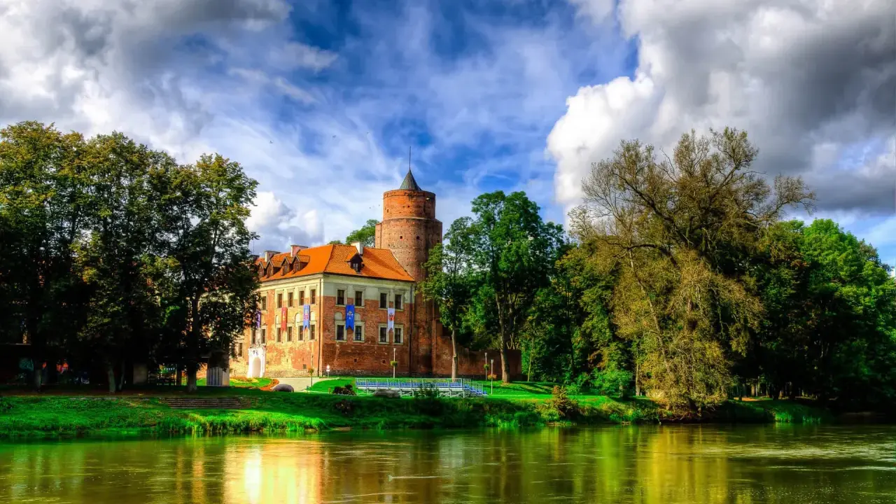 Malowniczy zamek w Łódzkiem nad rzeką, otoczony zielenią i chmurami.