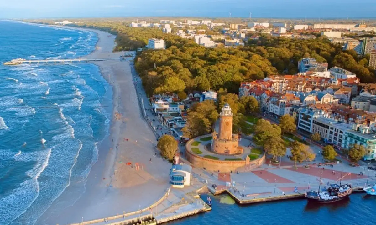 Malownicze miasta nad morzem z latarnią morską, plażą i molo. Widać port z żaglowcem i falujące morze.