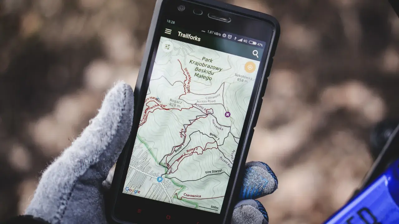 Rękawica trzyma smartfon z aplikacją **aplikacja trasy rowerowe** Trailforks, pokazującą mapę Beskidu Małego.