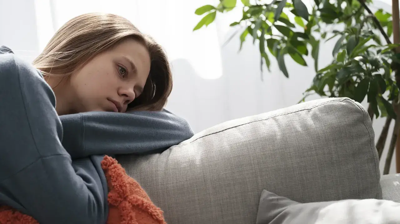En ung kvinna med utmattningssyndrom symtom vilar på en soffa, ser trött och nedstämd ut.