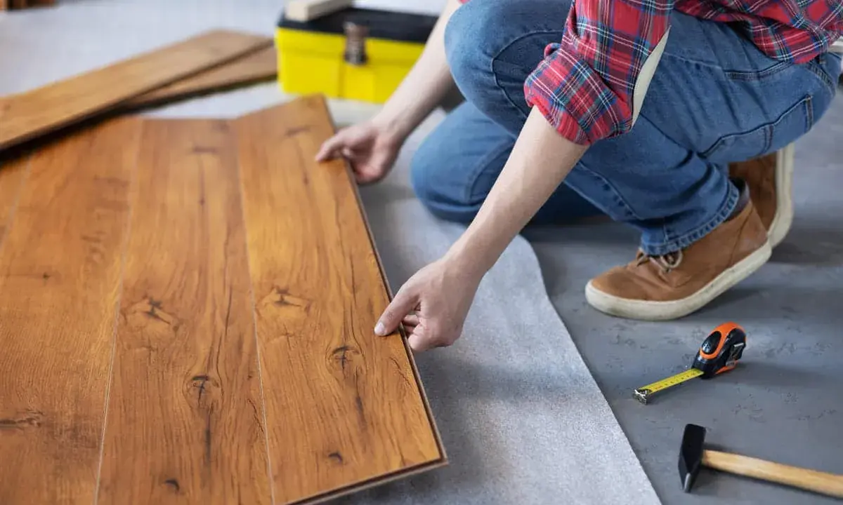 Persona instala tarima flotante, uniendo tablas de madera con cuidado. Herramientas como martillo y cinta métrica a mano.