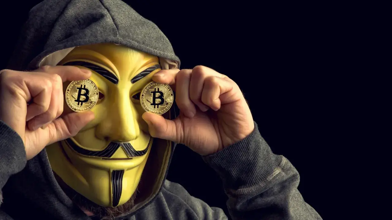 Osoba w masce Guya Fawkesa trzyma monety z Bitcoinem przed oczami. Czy MoonPay jest bezpieczny?