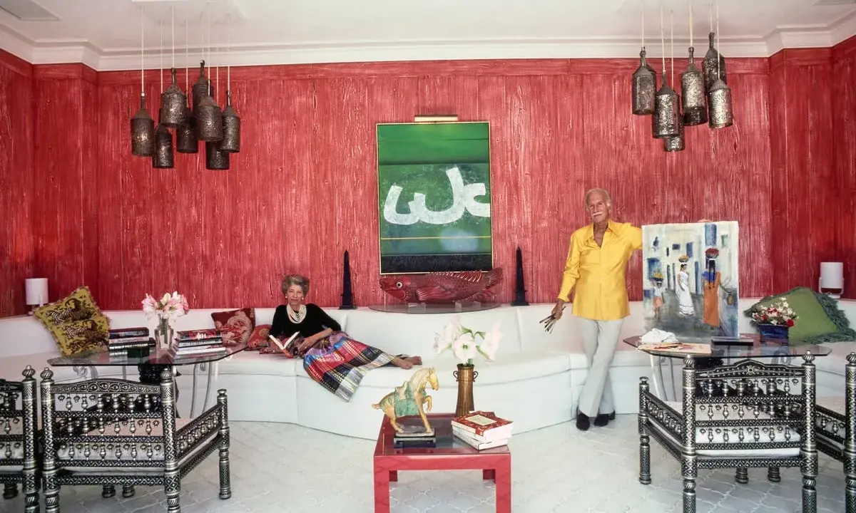 Interni di case anni '70: una donna legge sul divano bianco, un uomo mostra un quadro. Pareti rosse, lampade a grappolo.