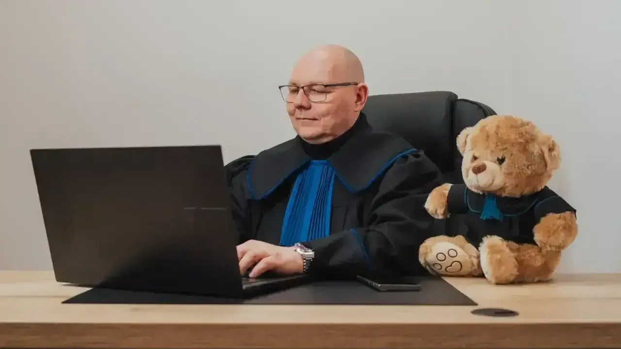 Prawnik w todze z pluszowym misiem obok, pracujący przy laptopie. Zastanawiasz się, ile kosztuje porada prawna?