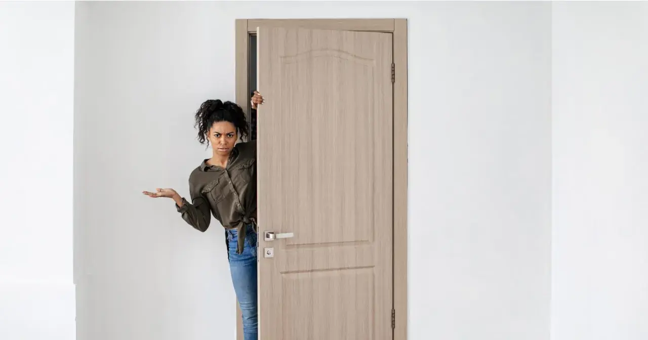 Mujer asomándose por una puerta de madera clara, con expresión de duda. Los colores de puertas como esta son muy versátiles.