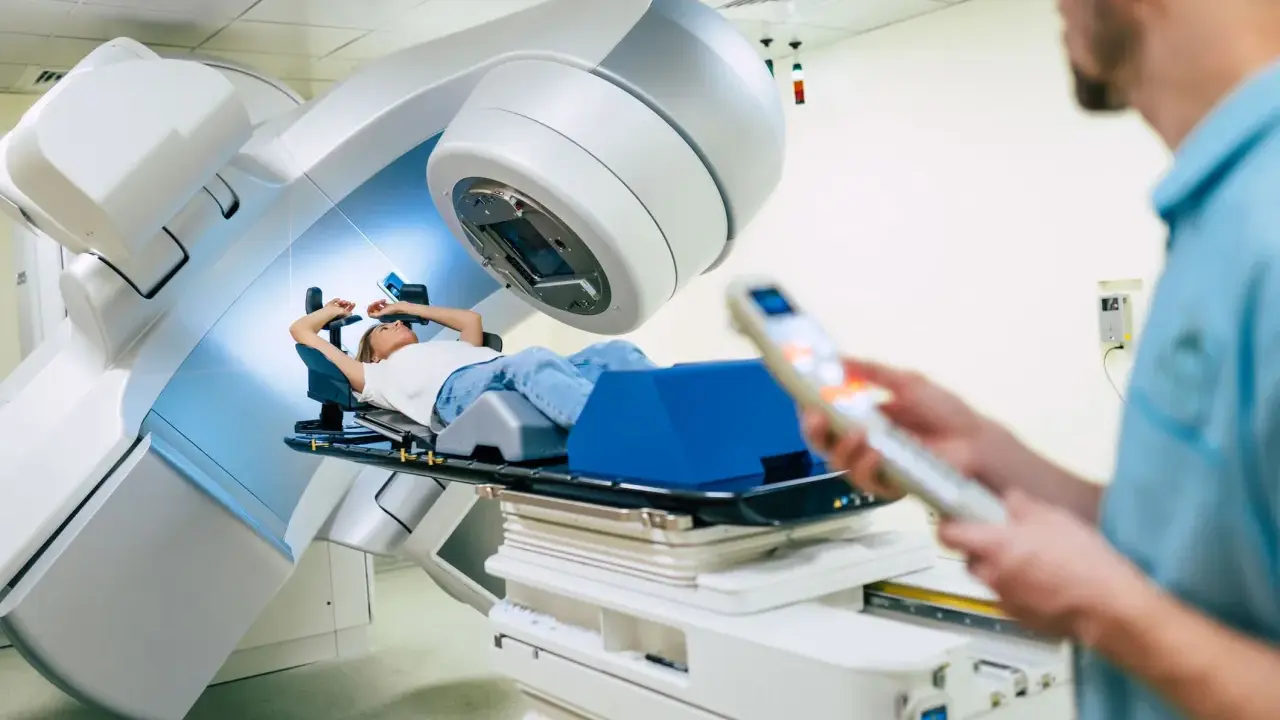 Ragazza sdraiata su lettino per radioterapia, tecnico in primo piano controlla il macchinario. Speranza di guarigione, ma con il timore dei radioterapia danni permanenti.