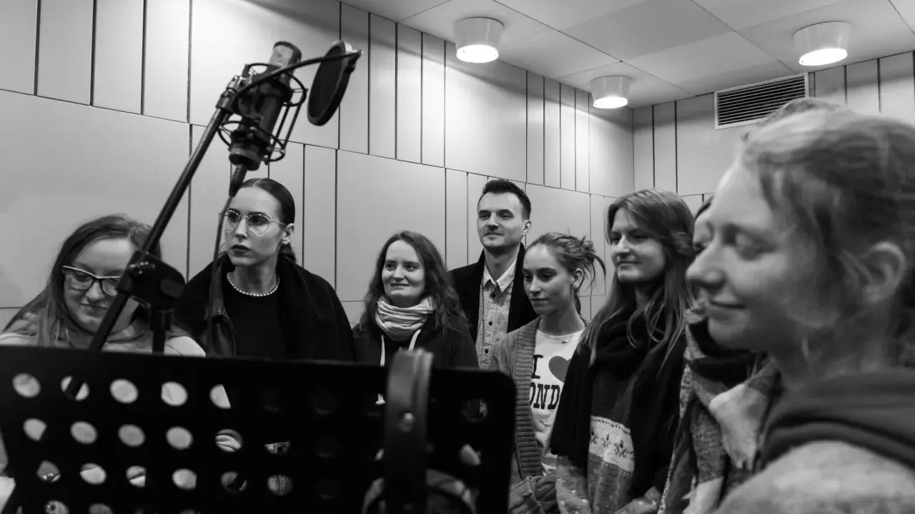 Grupa osób na kursie dubbingu, skupiona wokół mikrofonu w studiu nagraniowym.