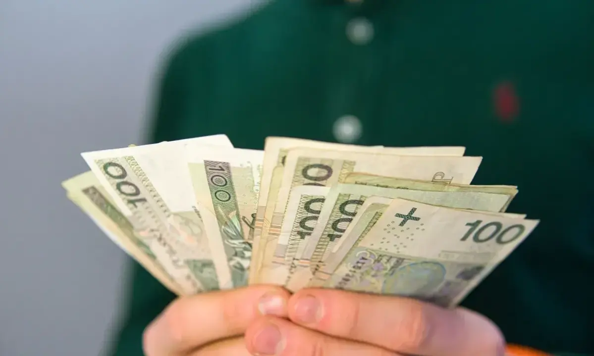 Dłonie trzymają wachlarz banknotów 100 zł. Może to być pomoc finansowa, np. renta po zmarłym ojcu, która zapewnia wsparcie.